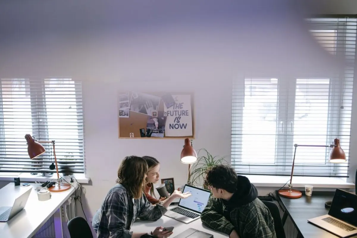 Two colleagues at a desk in a bright office talking over an open laptop, with a noticeboard on the wall behind them