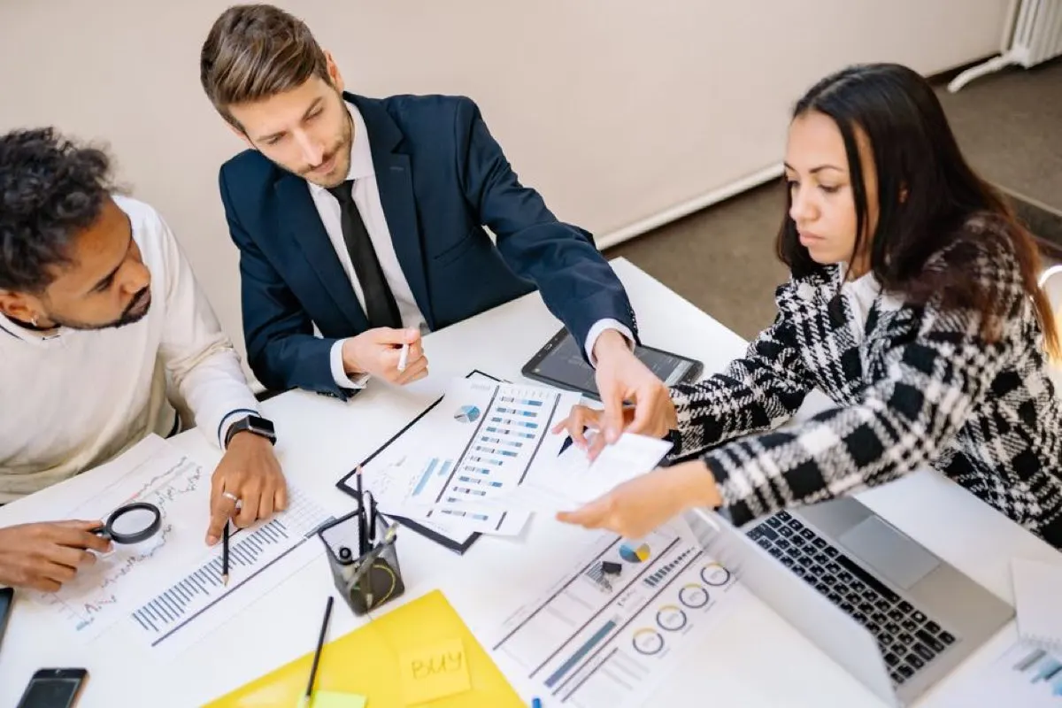 Three colleagues around a white desk reviewing printed charts and reports, one pointing at a figure on a page