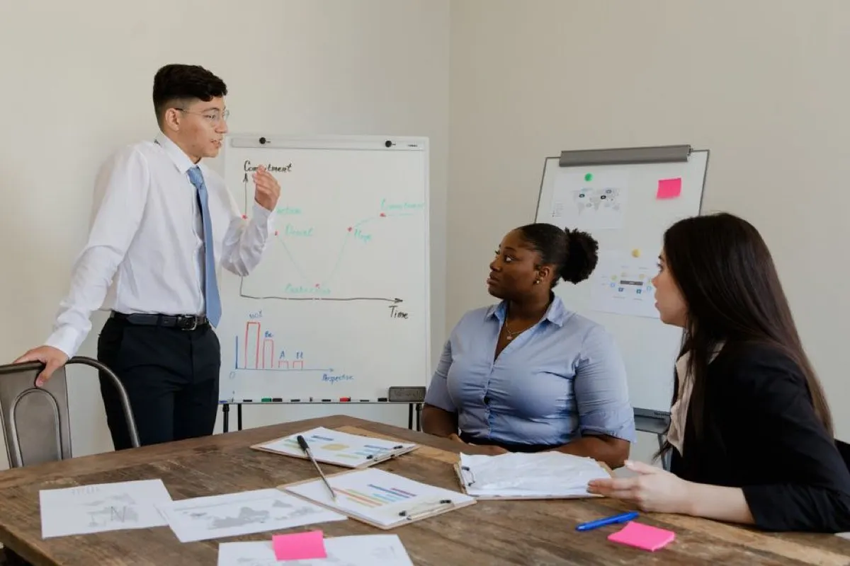 A man standing beside a flip chart with a hand-drawn line graph, presenting to two colleagues seated at a wooden table covered in printed charts