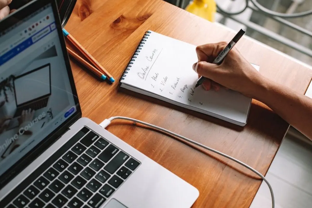 Overhead view of a laptop showing an online courses page beside a hand writing a numbered list headed Online Courses in a spiral notebook