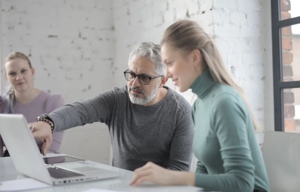 A man in glasses pointing at a laptop screen while a colleague beside him looks at it and a third person watches from behind