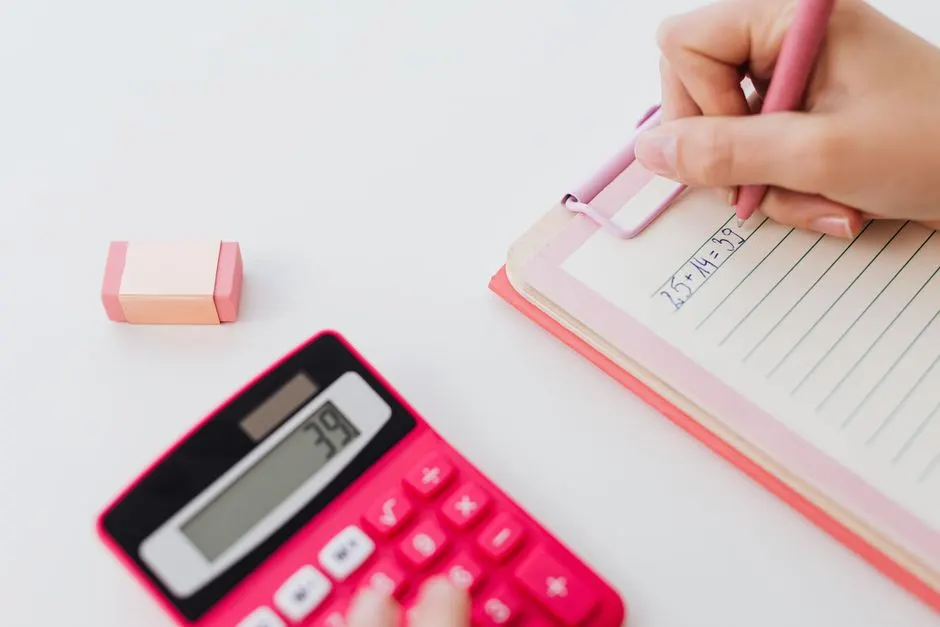 A hand writing a sum in a lined notebook beside a pink desk calculator and an eraser
