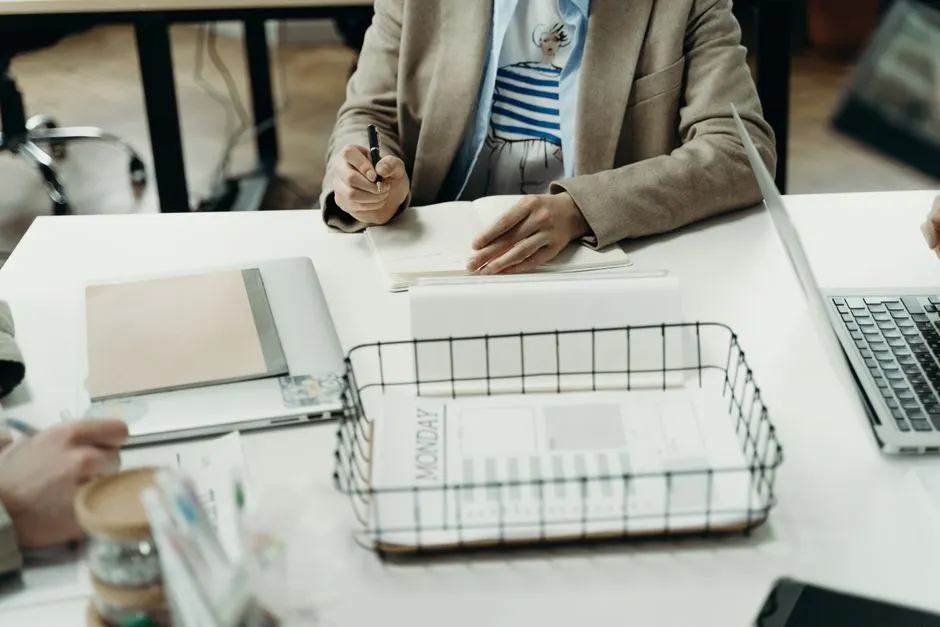 Someone writing in a notebook at a white desk with a laptop, a document tray and a printed weekly schedule headed Monday