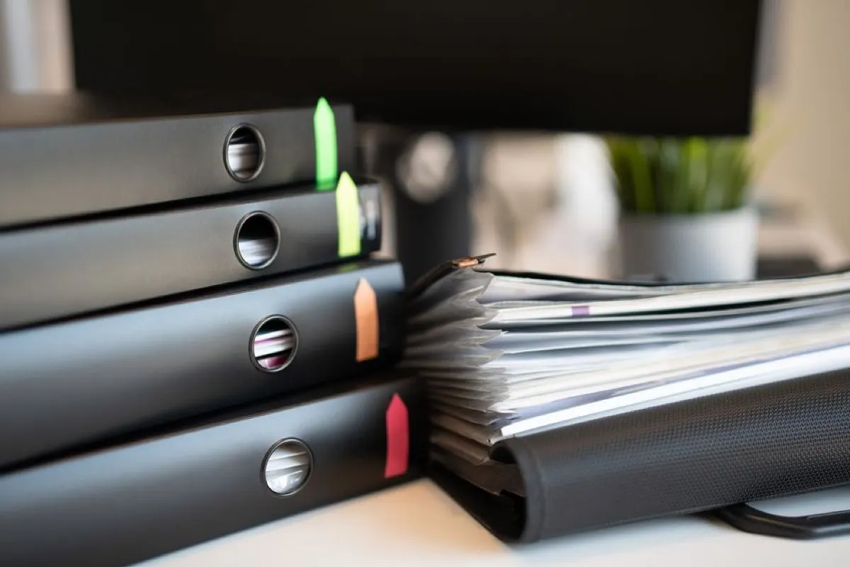 A stack of black lever-arch files with coloured index tabs beside a bulging document wallet on a desk