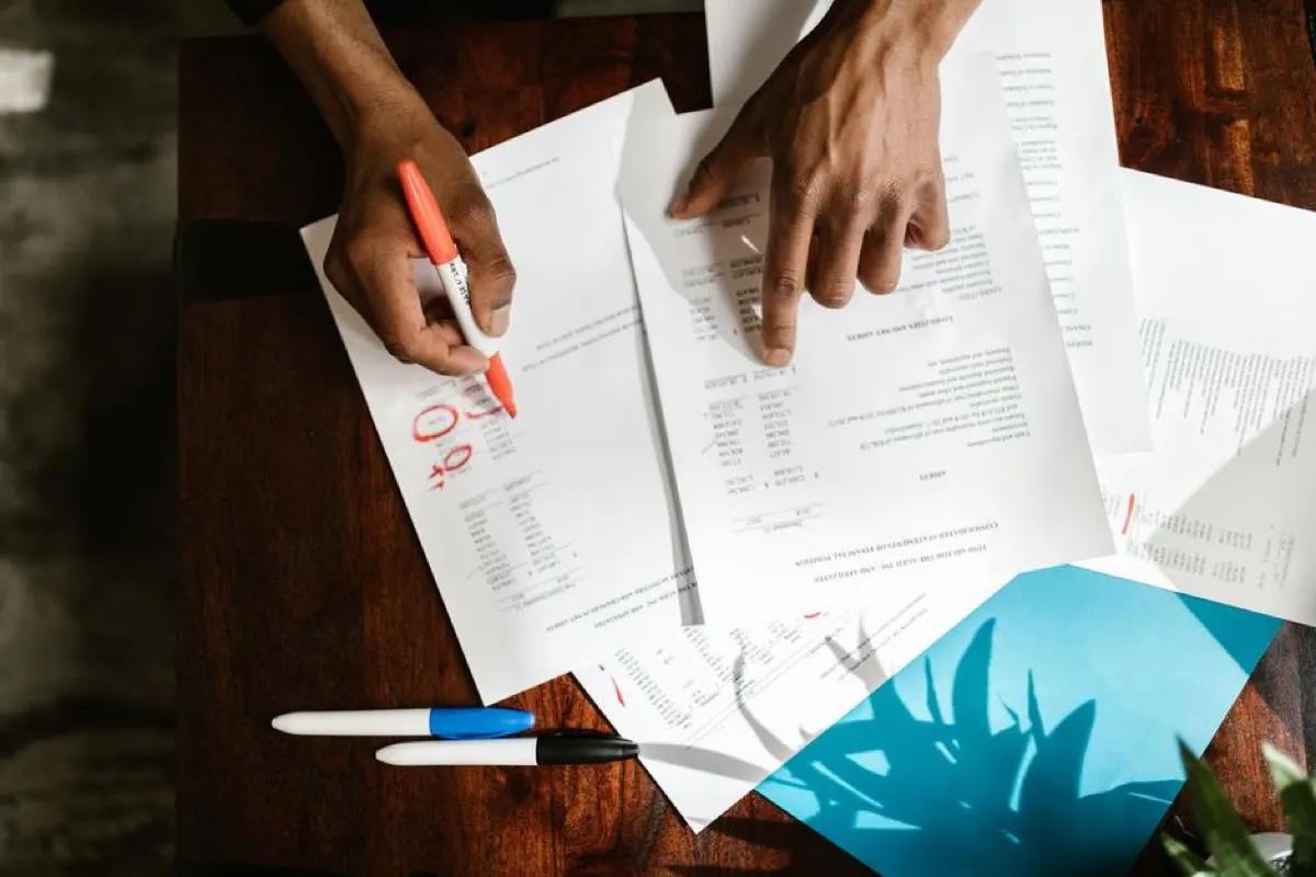 Overhead view of hands annotating printed financial documents on a dark wooden desk with marker pens