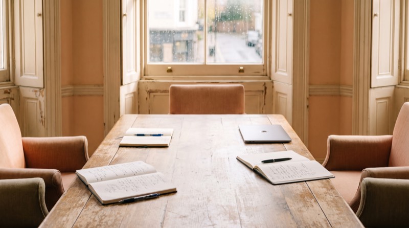 A meeting room table with open notebooks, pens and a laptop, lit by daylight