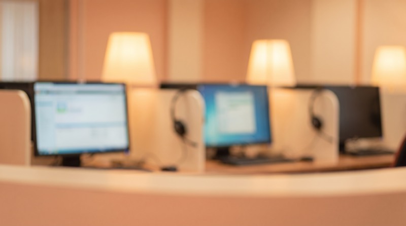 A row of contact centre desks with headsets and monitors showing support queues