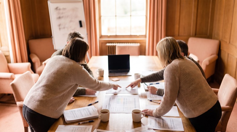 A meeting table with printed charts, a laptop and mugs of tea mid-discussion